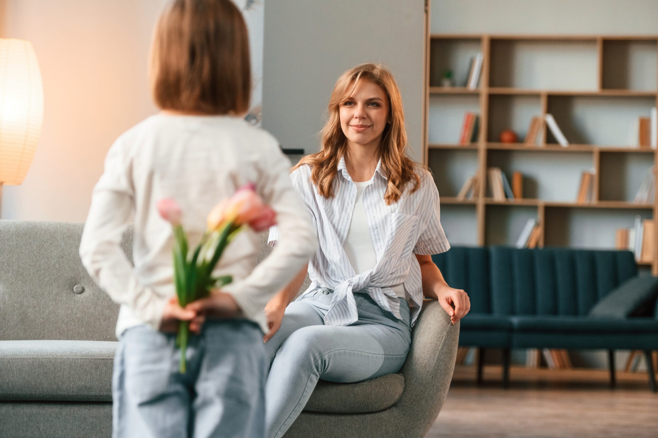 Girl is giving flowers for a woman. Mother with her little daughter is together in domestic room