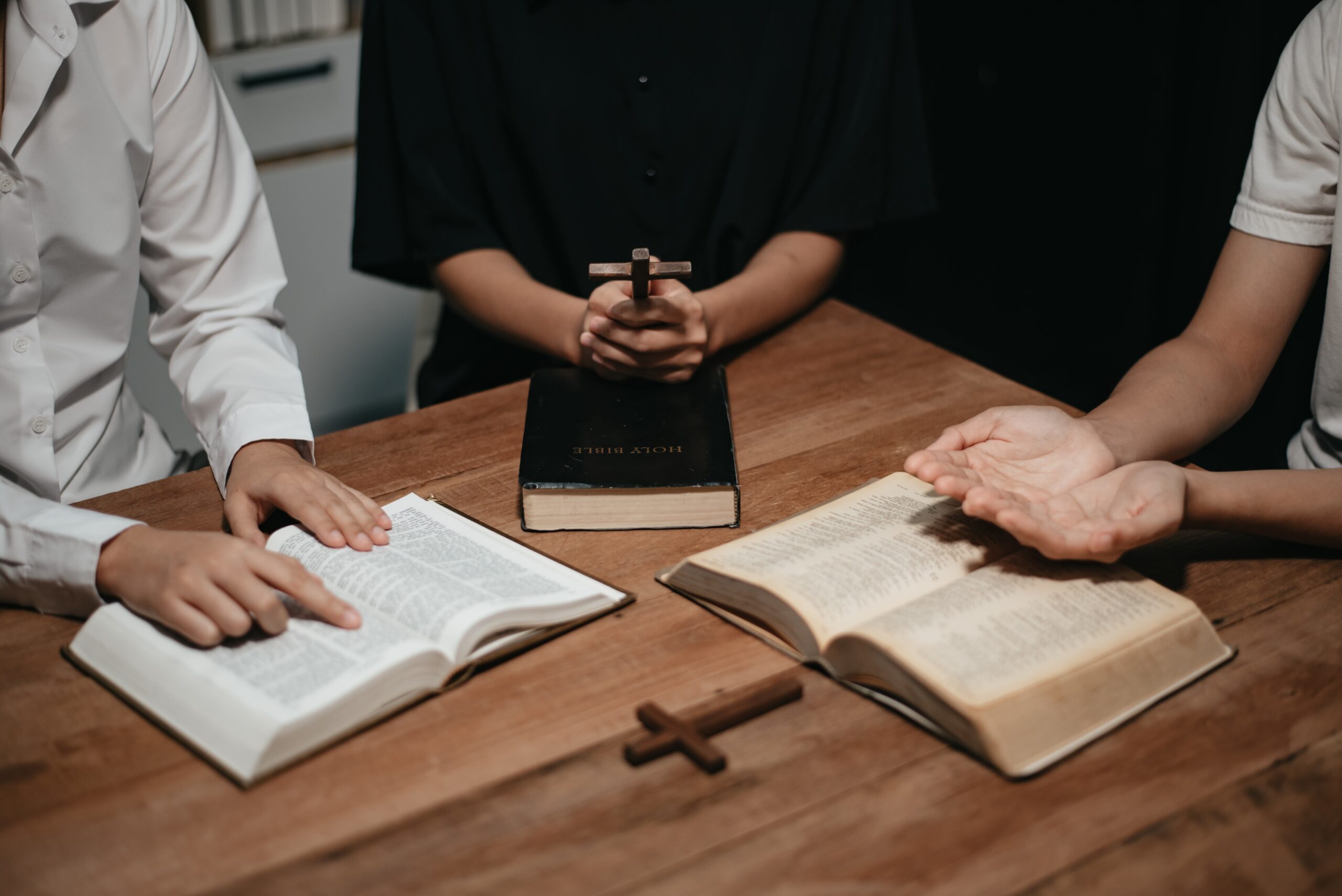 Group of Christians sit together and pray around a wooden table with blurred open Bible pages
