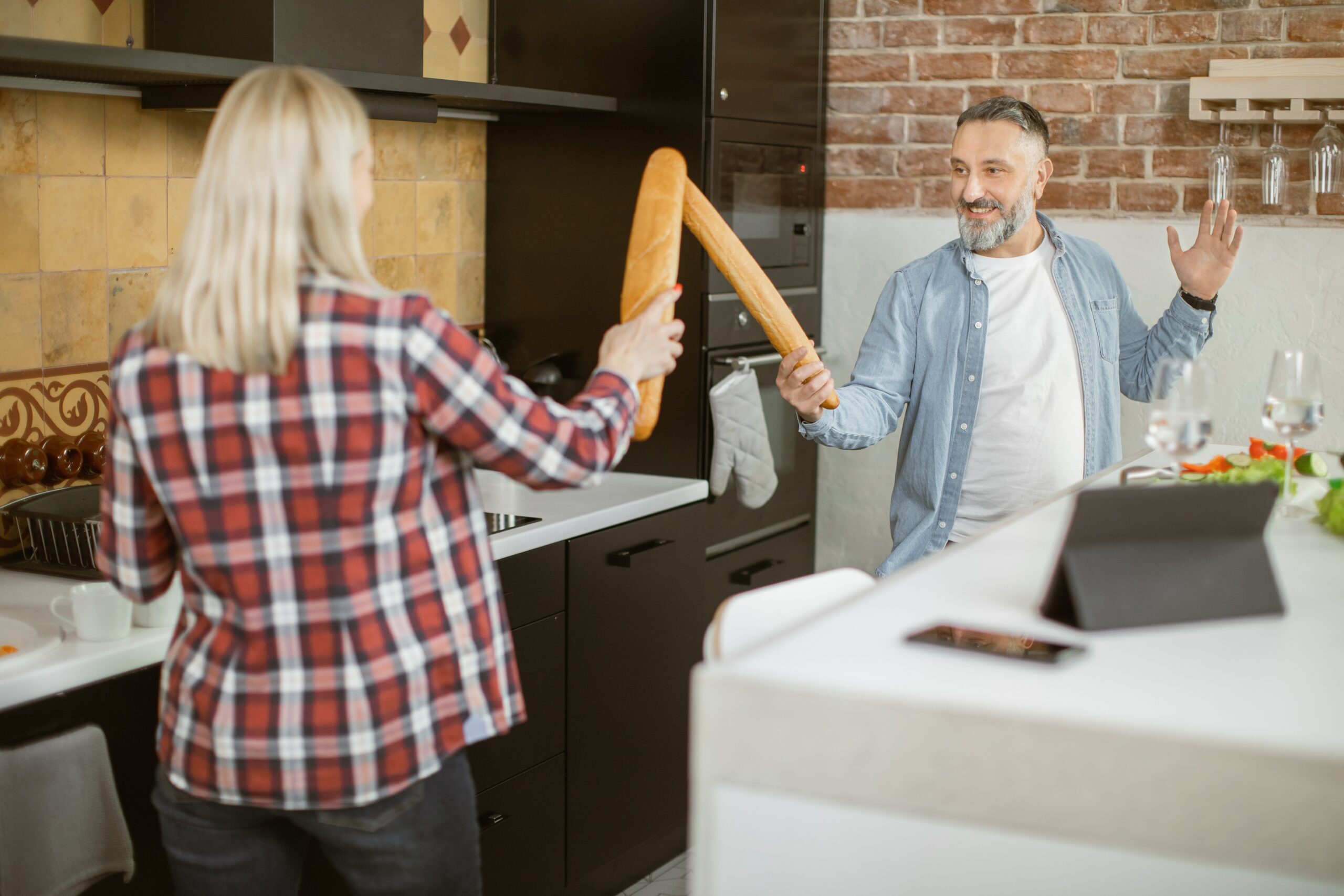 Joyful mature couple fighting with baguettes on kitchen