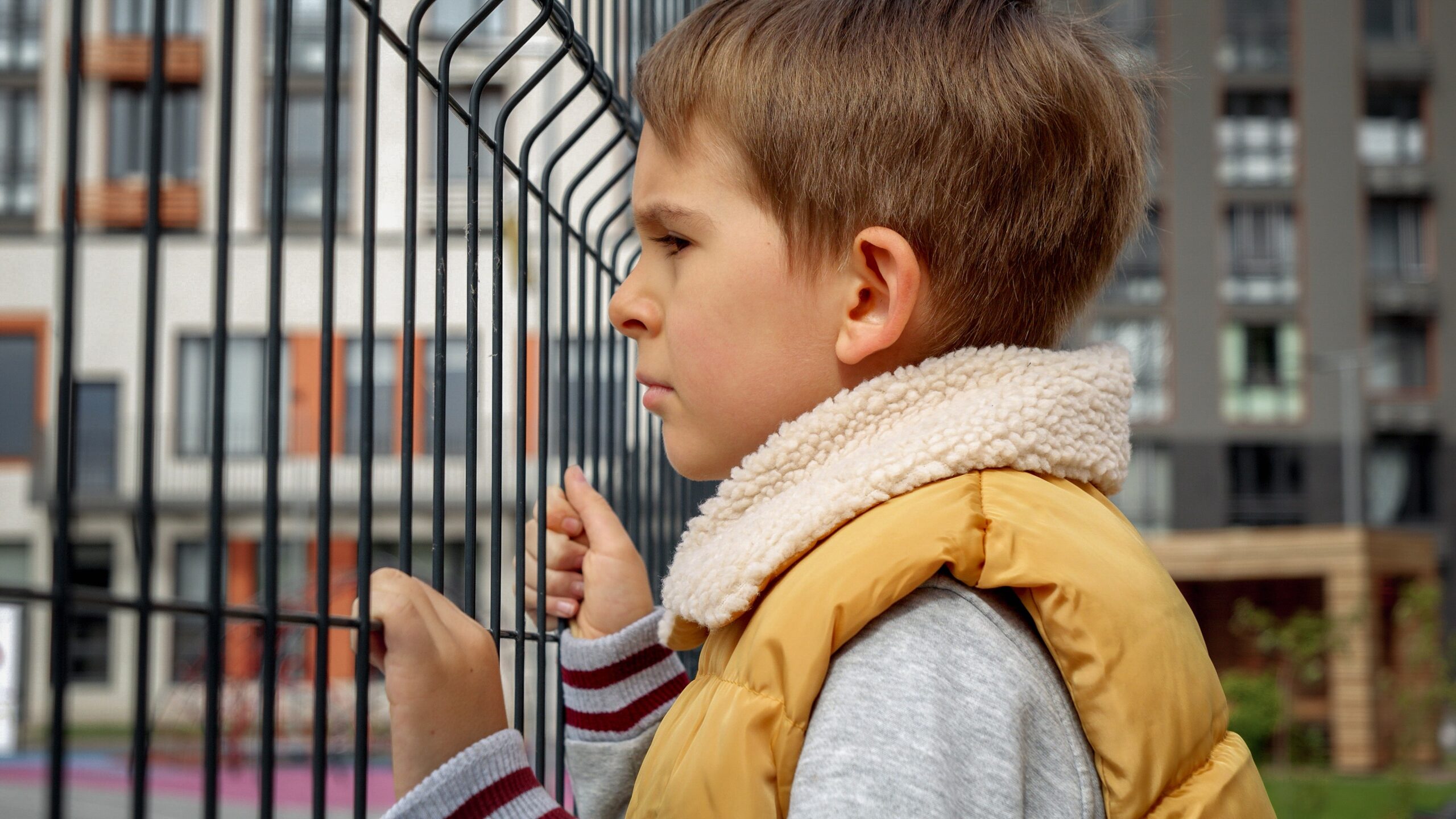 Portrait of sad little boy looking through metal fence