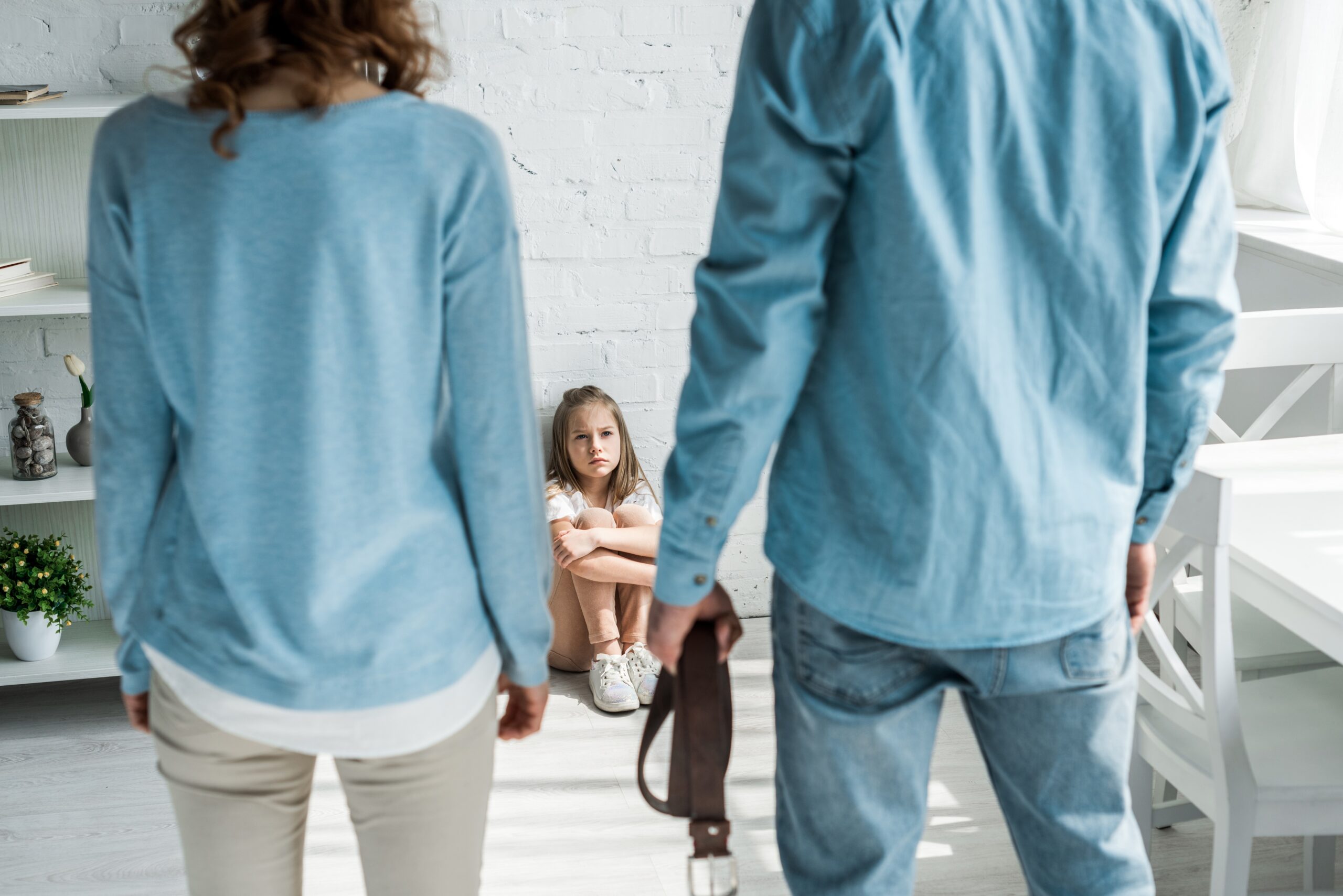 selective focus of scared kid sitting on floor and looking at father with belt standing near mother