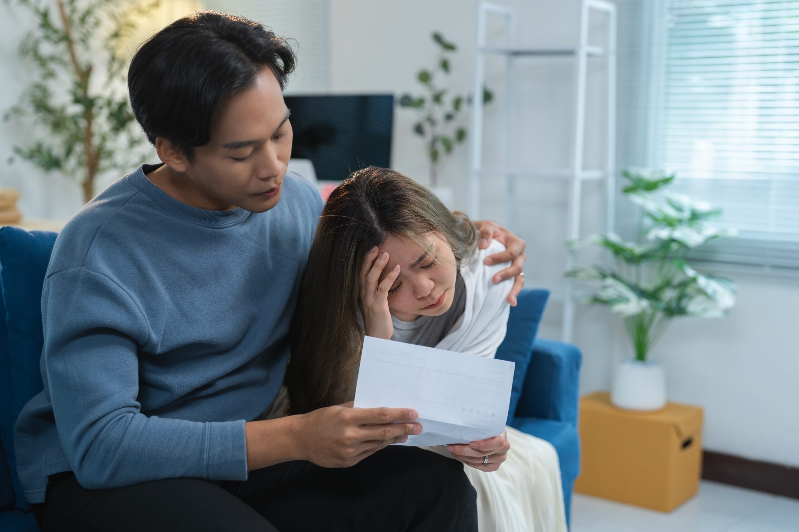 Stressed asian couple is reading a letter with bad news while sitting on the sofa at home, the woman